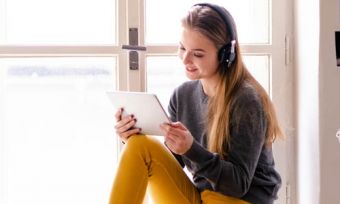 Young woman looking at tablet with headphones on, sitting at window