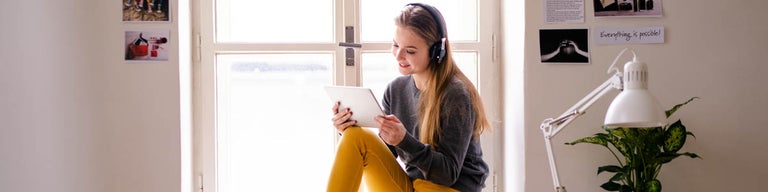Young woman looking at tablet with headphones on, sitting at window