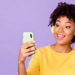 Young woman with headphones on looking at phone on purple background