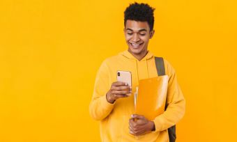 Young male student in yellow checking mobile phone happily