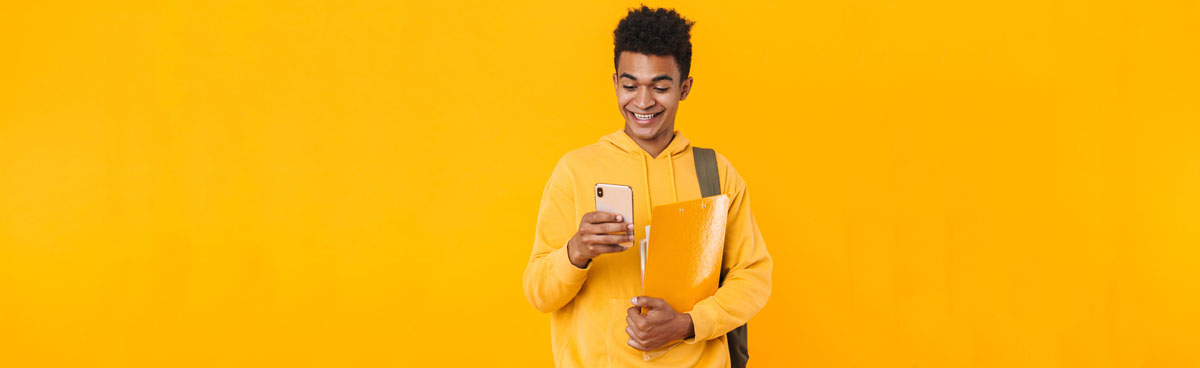 Young male student in yellow checking mobile phone happily