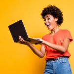 Young woman looking excitedly at computer against yellow background