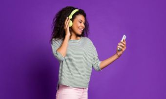 Young woman with headphones on looking at phone against purple background