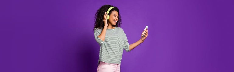 Young woman with headphones on looking at phone against purple background