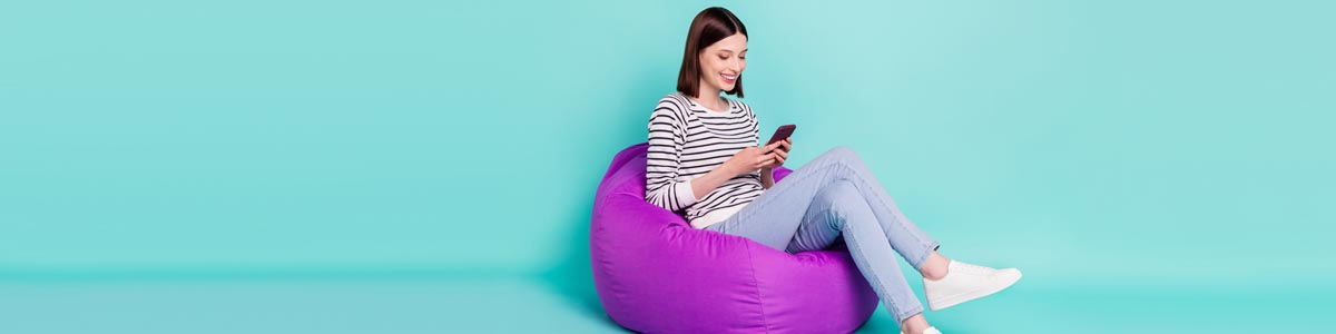 Woman looking at phone sitting on purple beanbag against teal background