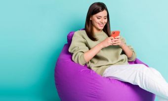 Young woman looking at mobile phone while sitting on beanbag against teal background