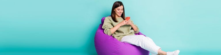Young woman looking at mobile phone while sitting on beanbag against teal background