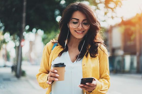 Young woman walking in city wearing headphones, looking and phone and holding coffee outdoors