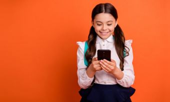 Girl in school uniform with backpack looking at phone against orange background