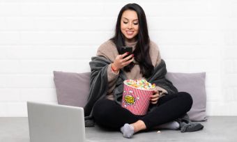 Young girl looking at phone while watching show on laptop and eating popcorn