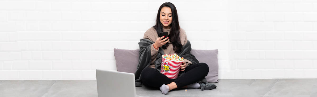 Young girl looking at phone while watching show on laptop and eating popcorn