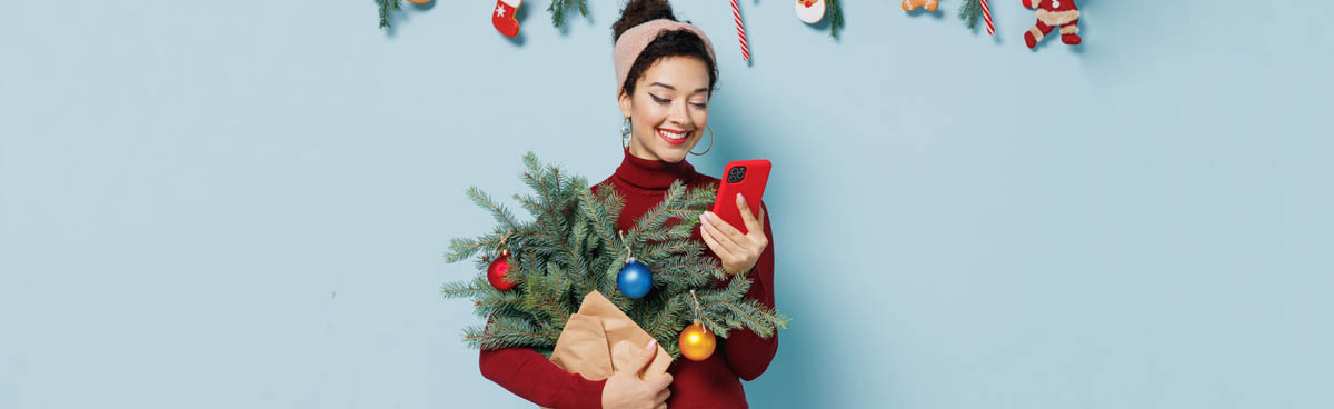 Smiling woman holding mini Christmas tree and looking at phone