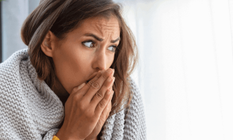 Woman warming her hands with her breath in front of a heater, wrapped in a blanket.