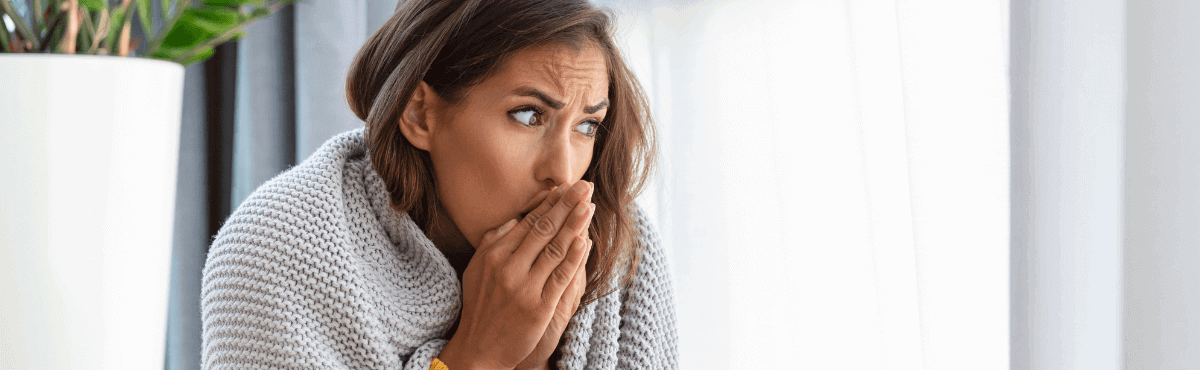 Woman warming her hands with her breath in front of a heater, wrapped in a blanket.