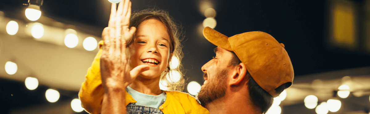 Father and child high-fiving in excitement, surrounded by outdoor lightbulbs.